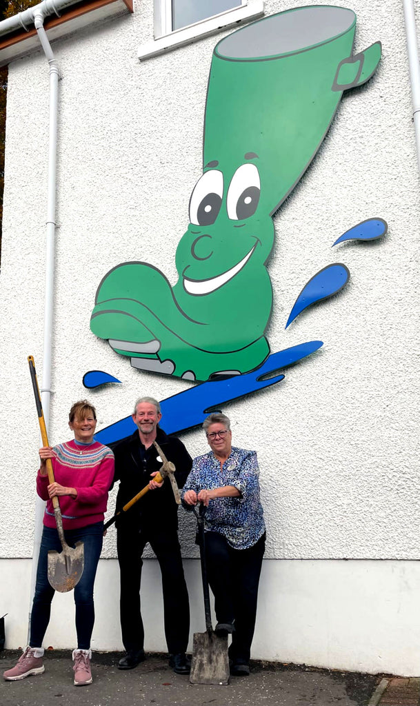 3 people, standing with shovels under a big green wellington boot sign with face