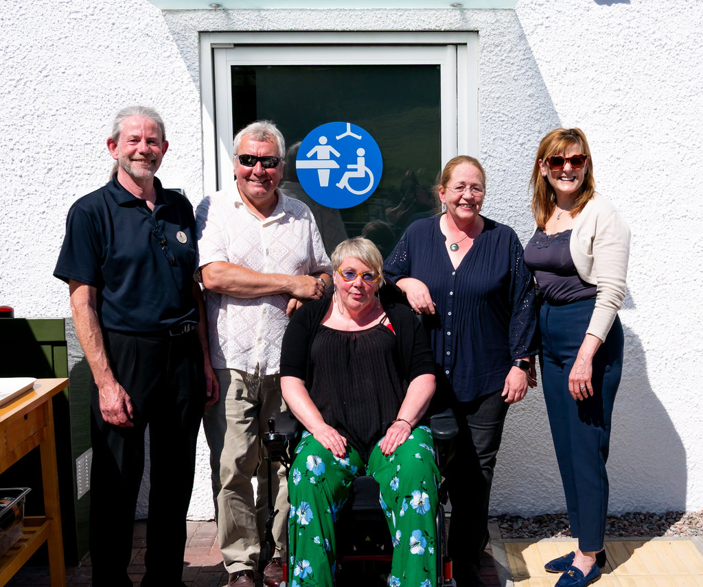 a group of 5 people standing in front of a door with changing places toilet logo, 2 men, 3 women - one on a wheelchair all dressed smart and middle aged