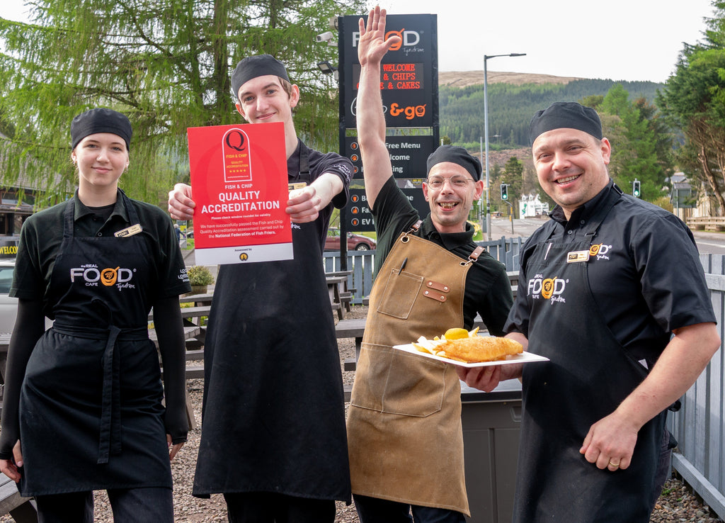 The Real Food cafe team dressed in uniforms holding fish n chips and NFFF quality accreditation are celebrating outside 