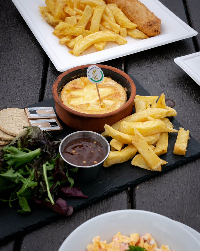 three appetising dishes on a table with a centre dishes served on a slate plate - it's a baked brie with a gluten free label on a toothpick served in a clay serving dishes next to chips, sweet chilli sauce, oatcakes, butter and some salad