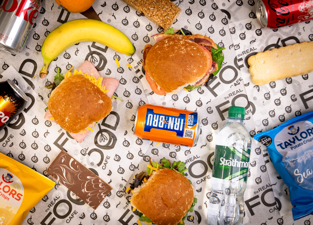 The Real food cafe packed lunches offering laid on a table including brown filled rolls, cans of soda (irnbru, cocacola_ flapjack, shortbread, millionaire shortbread, Taylors crisps of different flavours, banana, sparkling Strathmore water bottle