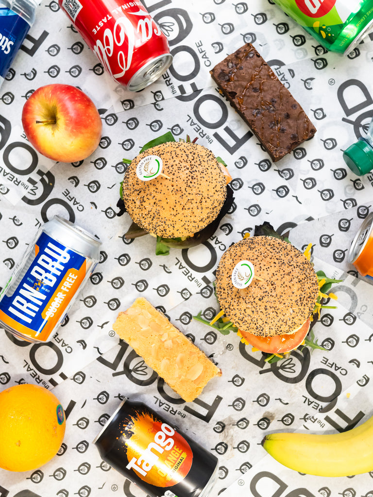 Gluten Free packed lunches offering from the real food cafe laid on a table - seeded filled rolls, chocolate brownie, Bakewell tart, tango, irnbru, apple and other drinks and fruits coming into the photo from the edges